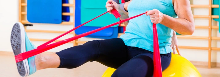 Person exercising on a yellow stability ball using a red resistance band, pulling the band while lifting one leg in a fitness studio