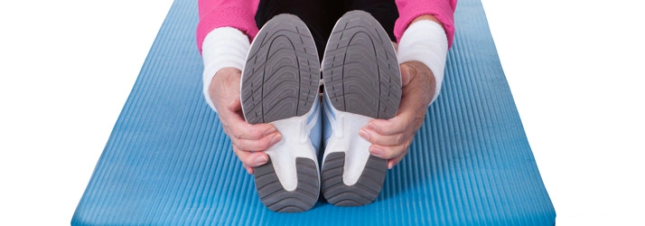 Person seated on a blue exercise mat, reaching forward to hold both feet in a stretching position