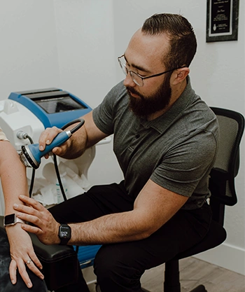 Jacob Covert performing a therapeutic procedure using a handheld device on another individual’s arm in a clinical setting, with medical equipment visible in the background